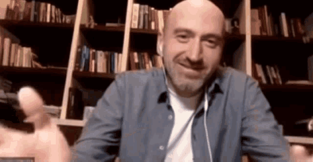 a bald man wearing ear buds is sitting in front of a bookshelf and smiling .