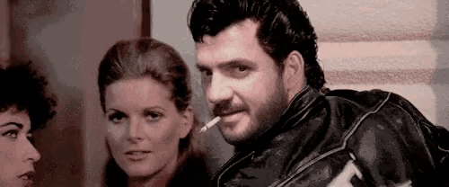 a man with a beard is smoking a cigarette while standing next to a woman .