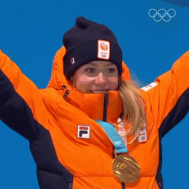 a woman wearing an orange jacket and a black beanie is holding up her arms and wearing a gold medal around her neck