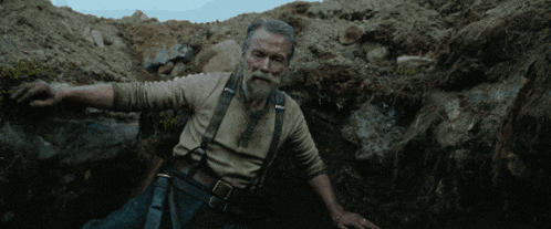 a man with a beard and suspenders is kneeling down in the dirt
