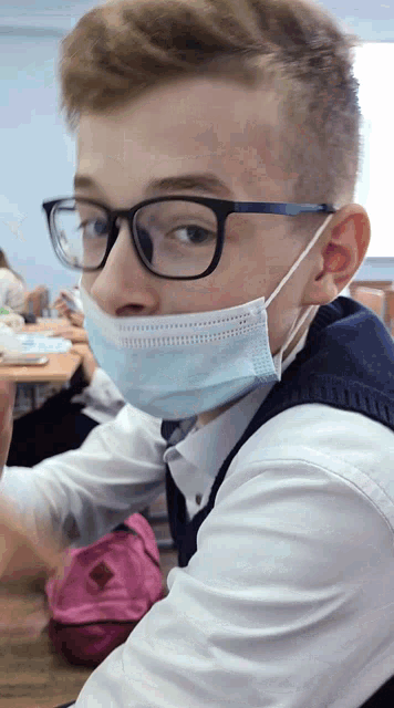 a boy wearing glasses and a face mask sits at a desk