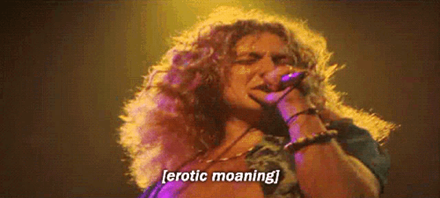 a woman with long curly hair is singing into a microphone and says erotic moaning
