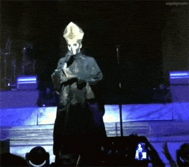 a man in a priest 's hat is standing on a stage holding a microphone .