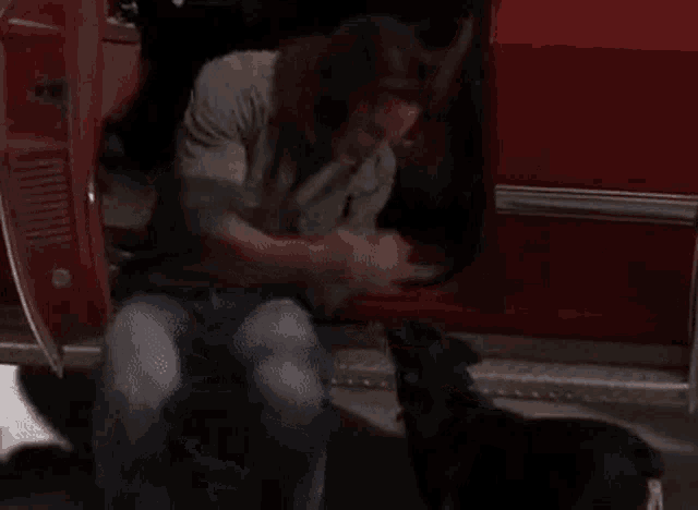 a man is kneeling down next to a black dog in the back of a red car .