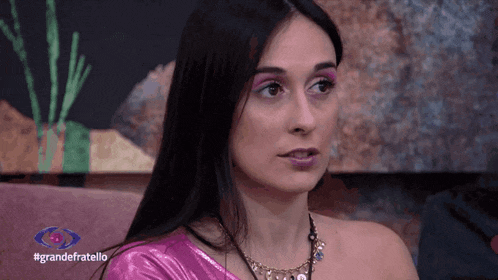a woman in a pink one shoulder top is sitting on a couch in front of a sign that says grande fratello