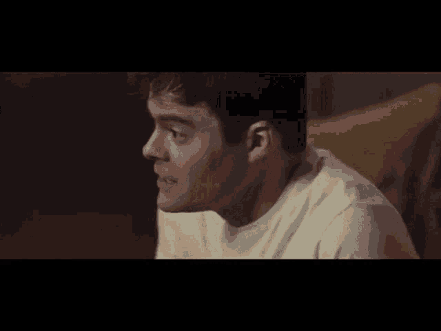 a man in a white shirt is sitting on a couch and looking at something .