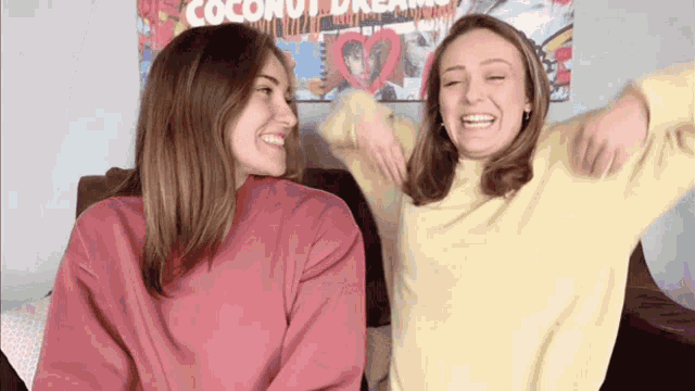 two women are sitting on a couch and laughing in front of a coconut dream poster .