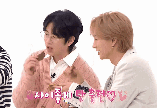 a man in a pink sweater is eating a chocolate bar next to a man in a white jacket