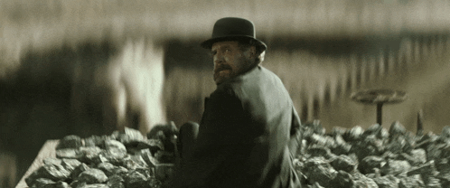 a man with a beard wearing a bowler hat is standing in a pile of rocks