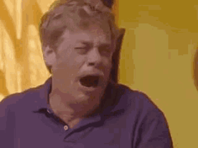 a man in a purple shirt is yawning with his mouth open and his eyes closed .
