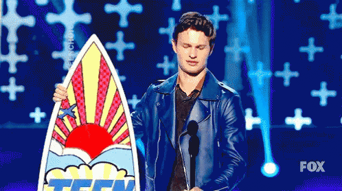 a man in a blue jacket is holding a surfboard on a stage at a fox awards show .