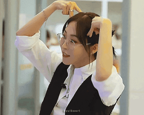 a woman wearing glasses and a vest is holding her hair in a ponytail .