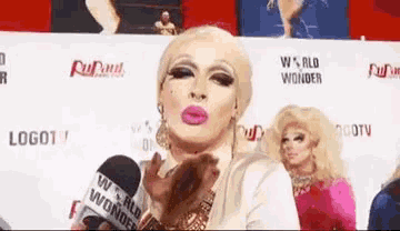 a drag queen is blowing a kiss while holding a microphone in front of a wall that says world wonder .