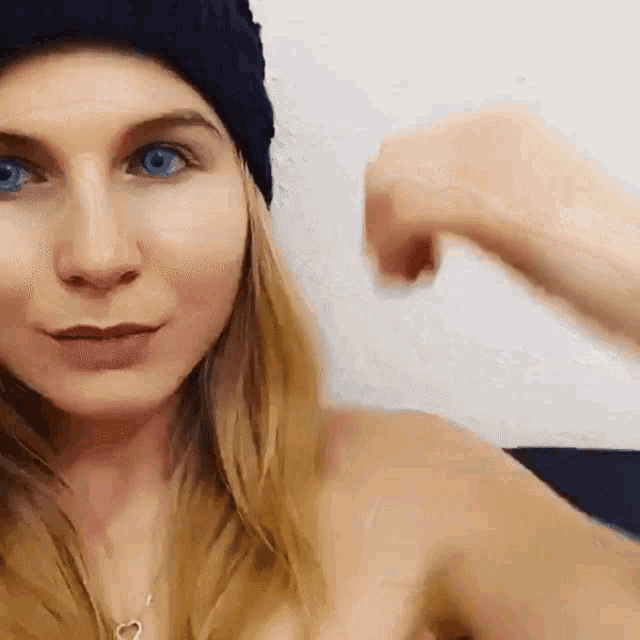 a woman wearing a blue hat and a heart necklace is flexing her muscles