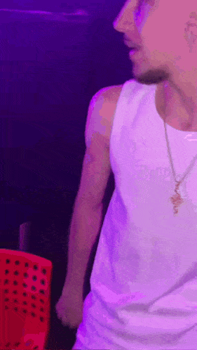 a man wearing a white tank top and a necklace is standing in front of a purple background .