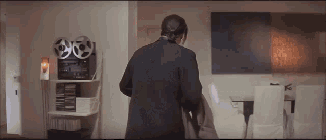 a woman in a black jacket is standing in a living room with a tape recorder in the background