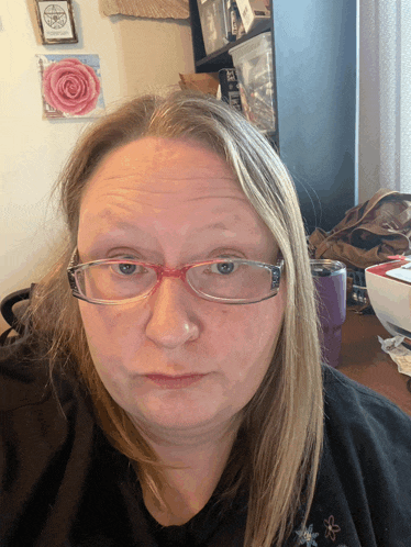 a woman wearing glasses looks at the camera with a pentagram hanging on the wall behind her