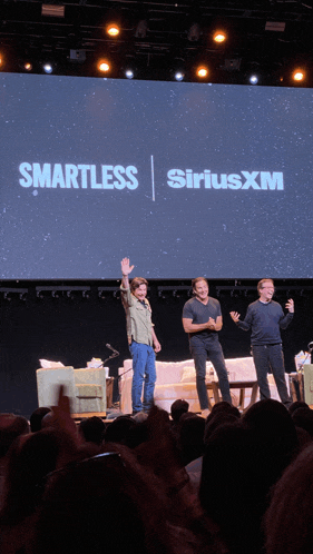 three men are standing on a stage in front of a large screen that says smartless siriusxm