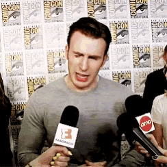a man is talking into a microphone with the letter b on it in front of a comic con wall
