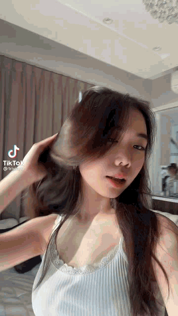 a woman in a white tank top is standing in front of a mirror and holding her hair .