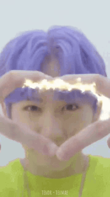 a person with purple hair is making a heart shape with their hands .