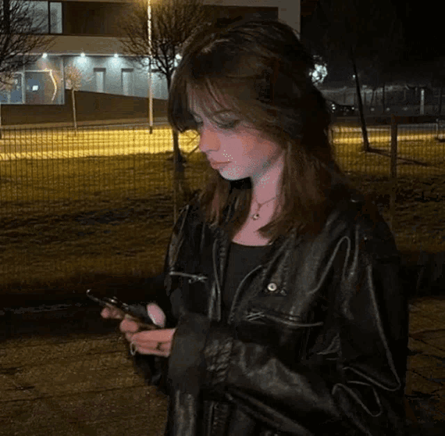 a young woman wearing a black leather jacket is standing in front of a building at night .
