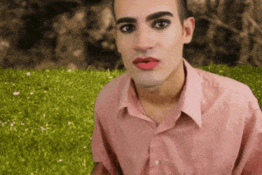 a man wearing a pink shirt and red lipstick is sitting in the grass