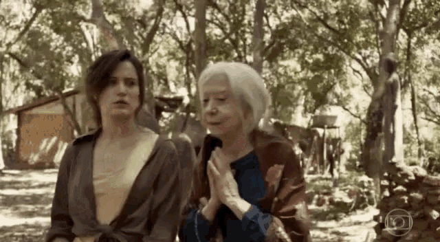 two women are standing next to each other in a forest with their hands folded in prayer .