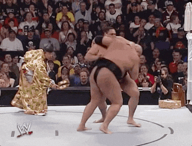 two sumo wrestlers are fighting in front of a crowd with a w logo on the ground .