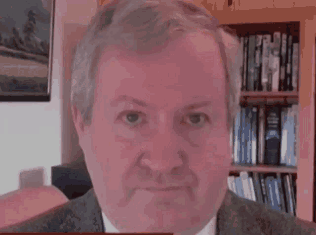 a man in a suit and tie is looking at the camera with a bookshelf in the background
