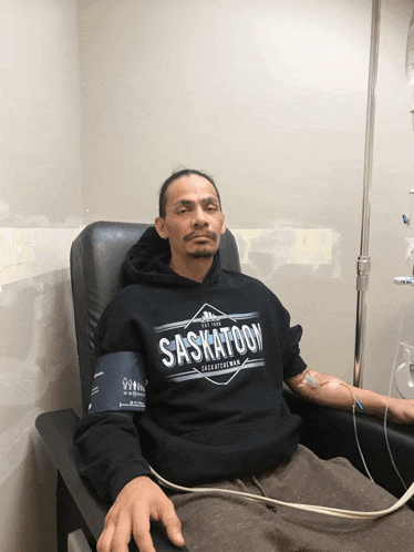 a man wearing a saskatoon sweatshirt sits in a chair