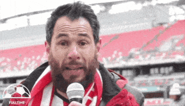 a man with a beard is holding a microphone in front of a soccer field that says fulltime
