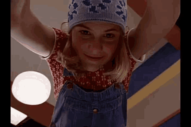 a young girl wearing overalls and a beanie is hanging upside down in a room .