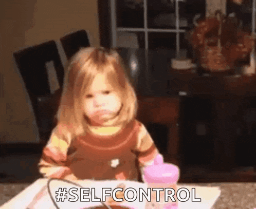 a little girl is sitting at a table with a pink cup and a spoon and making a face .