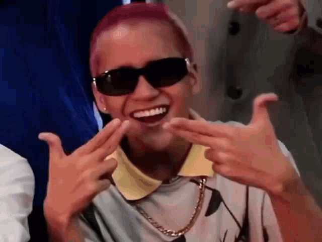 a young man with pink hair is wearing sunglasses and making a peace sign with his hands .