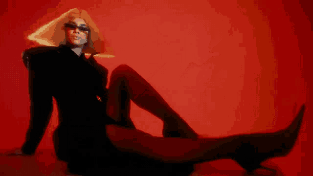 a woman in a black dress and sunglasses is laying on the floor in front of a red background