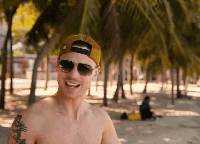 a shirtless man wearing sunglasses and a hat smiles