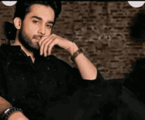 a man with a beard is sitting on a couch with his hand on his chin and wearing a watch .
