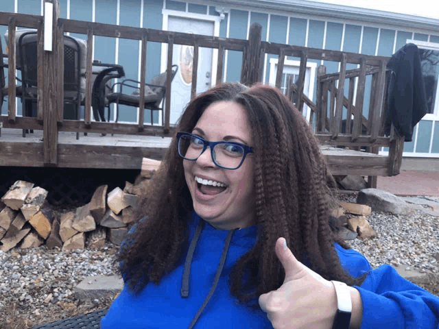 a woman wearing glasses and a blue sweatshirt gives a thumbs up