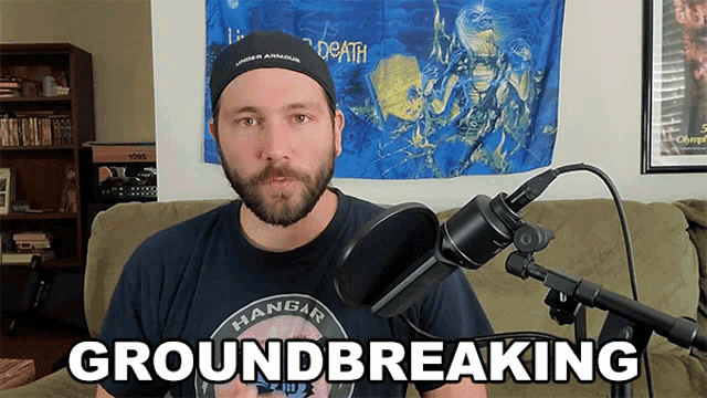 a man with a beard stands in front of a microphone with the words groundbreaking written below him