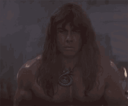 a shirtless man with long hair and a necklace around his neck is looking at the camera .