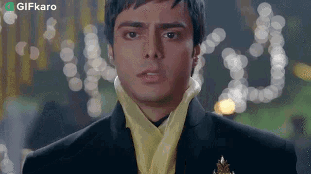 a man wearing a black suit and a yellow scarf looks at the camera with a gifkaro logo in the corner