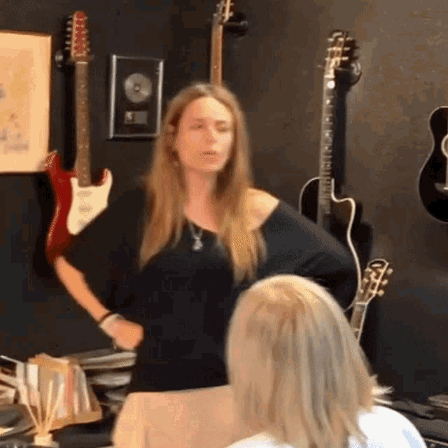 a woman in a black shirt stands in front of a wall of guitars