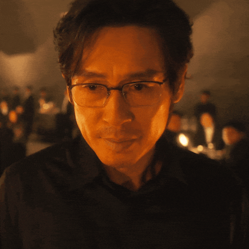 a man wearing glasses and a black shirt looks at the camera in a dark room