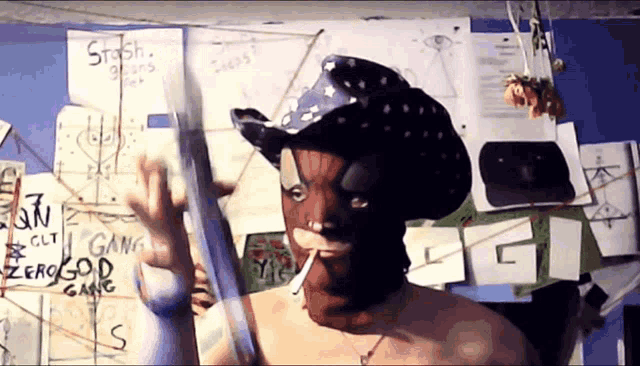 a man wearing a cowboy hat is smoking a cigarette in front of a board that says stash on it