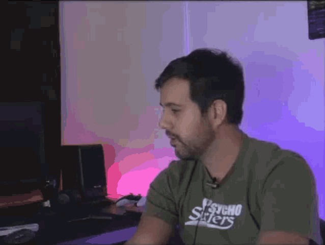 a man wearing a psycho surfers t-shirt is sitting at a desk in front of a computer .