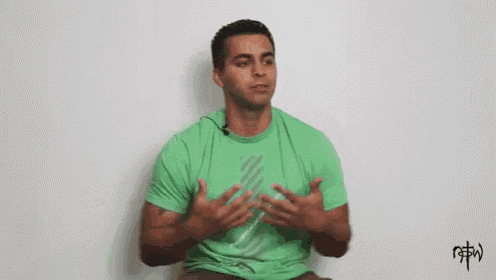 a man in a green t-shirt is sitting in front of a white wall with his hands on his chest .