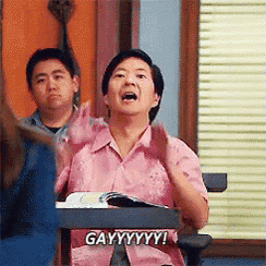 a man in a pink shirt is sitting at a desk that says gayyyyy