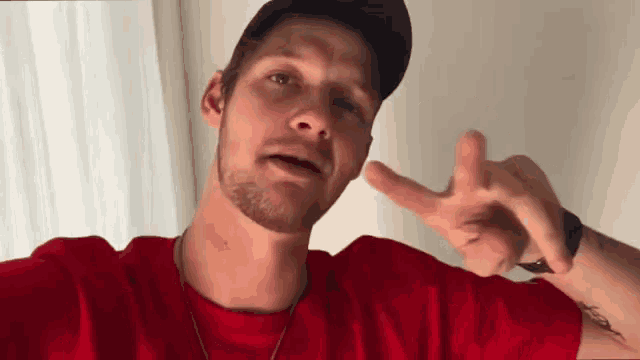 a man wearing a red t-shirt and a black hat is giving the middle finger