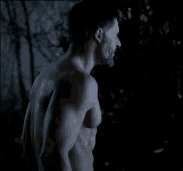 a shirtless man with a beard is standing in the dark in a dark forest .